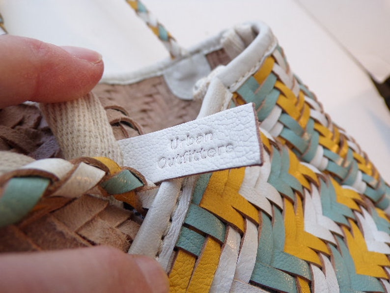 May include: White leather sandals with a woven design in yellow, blue, and white. The sandals have a white leather label that says "Urban Outfitters".