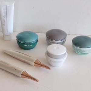 Könnte beinhalten: Eine Auswahl an Hautpflegeprodukten, darunter Tuben und Tiegel. Die Produkte sind weiß, türkis und grau, mit goldfarbenen Akzenten. Der Markenname "ANEW CLINICAL" ist auf einer Tube sichtbar.