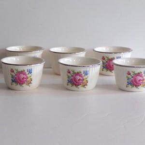 Könnte beinhalten: Sechs cremefarbene Keramikschalen mit silbernem Rand und einem Kreuzstich-Blumendesign mit rosa Rosen und blauen Akzenten. Die Schalen sind auf einer weißen Oberfläche angeordnet.