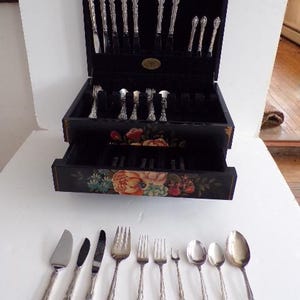 May include: A silver-toned cutlery set presented in a black wooden case with a floral design. The set includes knives, forks, spoons, and serving utensils. The case is open, displaying the neatly arranged silverware.