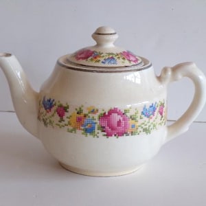 May include: Cream-colored ceramic teapot with a floral cross-stitch design around the body and on the lid. The teapot has a curved spout and a curved handle. The floral design includes pink, blue, yellow, and green flowers.