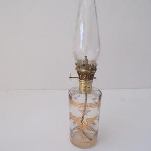 May include: A clear glass oil lamp with a gold-coloured design of two birds with outstretched wings. The lamp has a clear glass chimney and a gold-coloured metal base.