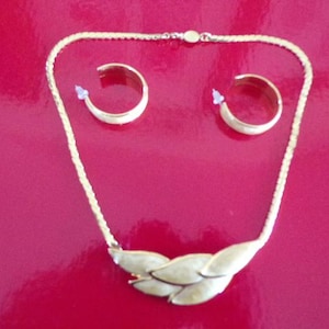 Puede incluir: Un collar de cadena dorada con un colgante blanco en forma de hoja. El collar viene con un par de pendientes de aro dorados a juego.