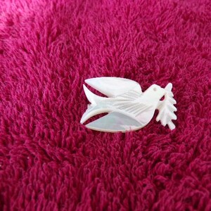 May include: A white dove brooch with iridescent detailing, carrying an olive branch. The brooch is set against a textured, deep pink background. The dove's wings are spread. A symbol of peace and hope.