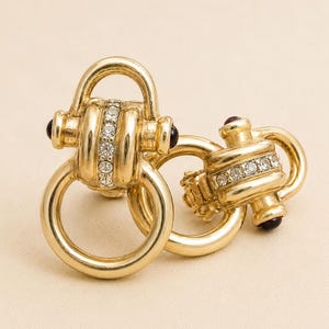 May include: A pair of gold-toned metal earrings featuring a circular hoop design connected to a central bar embellished with a row of clear, sparkling rhinestones. Each earring is accented with small, dark, rounded end caps on the sides.