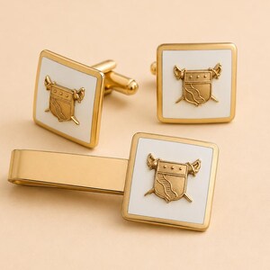 May include: A set of gold-toned metal accessories including two square cufflinks and one tie bar. Each piece features a white square inlay centered with a raised gold-toned crest design depicting a shield flanked by two crossed swords.