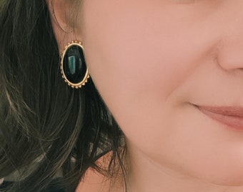 May include: A close-up of a woman's face wearing a pair of gold oval earrings with black stones. The earrings have a beaded edge.