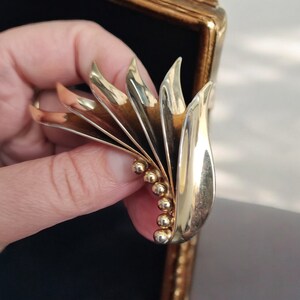 May include: A gold-toned brooch with a stylized leaf design. The brooch is made of metal and has a series of small, round beads along the bottom edge.