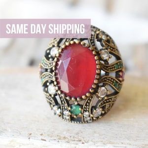 May include: A large, ornate ring with a red gemstone surrounded by smaller clear and green gemstones. The ring is made of gold-toned metal.