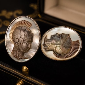 May include: Two oval-shaped cufflinks with detailed relief carvings. One features a profile of a figure in a helmet, the other a profile of a figure with a headdress. Both are set in a silver-toned metal with a mother-of-pearl background, displayed in a black box.