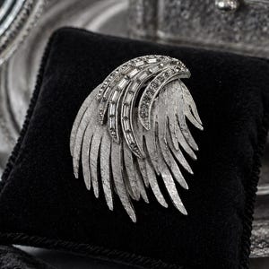 Puede incluir: Broche plateado con forma de ala estilizada, adornada con cristales transparentes y ahumados. La broche se exhibe sobre un cojín de terciopelo negro, con un espejo y una caja decorativa en el fondo.