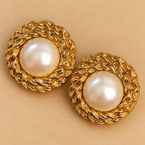 May include: A pair of round earrings featuring a large, smooth, cream-colored faux pearl at the center. The pearl is surrounded by a thick, textured gold-tone metal border designed with a twisted rope pattern that encircles the entire circumference.
