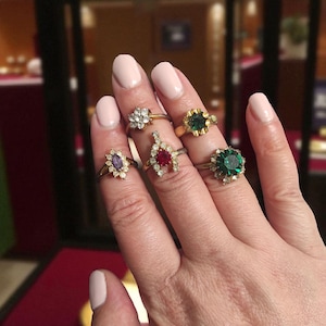 Puede incluir: Una mano que muestra seis anillos con varios engastes de piedras preciosas. Los anillos tienen bandas de oro e incluyen un anillo en forma de flor con piedras transparentes, un anillo con una piedra roja y un anillo con una piedra verde grande.