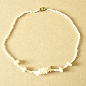 May include: A delicate, off-white necklace featuring small, round beads and larger, irregular-shaped beads. The necklace is secured with a gold-colored clasp. The design is simple and elegant, suitable for various occasions.