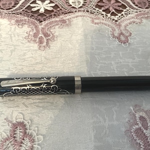May include: A black fountain pen featuring a silver-toned metal clip and a decorative silver filigree pattern on the cap. The pen has a smooth, cylindrical barrel and a polished finish, with a silver-colored band separating the cap from the body.