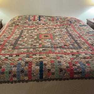 May include: A large quilt featuring a multi-colored yo-yo pattern constructed from numerous small, circular fabric pieces. The design includes a central square, concentric rectangular borders, and a scalloped edge, showcasing a variety of floral and solid-colored textile scraps in shades of pink, blue, yellow, and green.