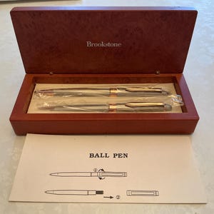 May include: A Brookstone branded wooden presentation box containing two silver and gold-tone ballpoint pens wrapped in plastic. Below the box is an instruction card with the text BALL PEN and diagrams showing how to operate the pen mechanism.
