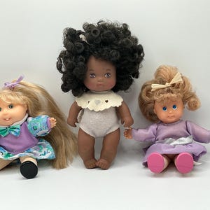 May include: Three dolls with distinct features. The left doll has blonde hair, a purple and floral dress, and a lavender bow. The center doll has dark curly hair, a white scalloped collar, and a light gray bodysuit. The right doll has light brown hair, a lavender dress, and a cream bow.
