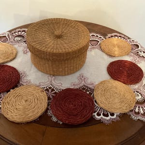May include: A round, lidded woven basket in a natural tan color, accompanied by six circular woven coasters. Three of the coasters are a natural tan color, and three are a deep reddish-brown color, all featuring a spiral-patterned texture.