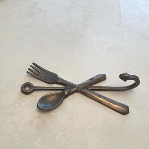 May include: A dark gray metal wall hook featuring a decorative design of a crossed fork and spoon. The fork and spoon are joined at the center, with the fork handle extending upward into a curved hook shape and the spoon handle extending downward.