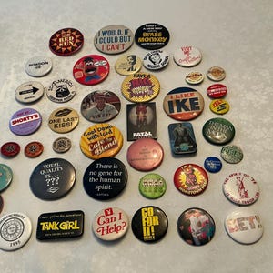 May include: A collection of assorted circular and rectangular pinback buttons featuring various colors, graphic designs, and text including phrases like I Would If I Could But I Can't, I Like Ike, Tank Girl, Can I Help, and Go For It, arranged in a group.
