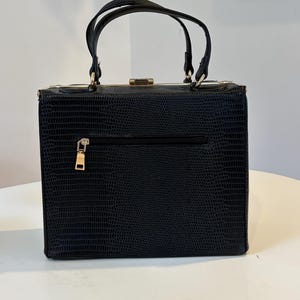 May include: A black, structured handbag with a textured, reptile-like pattern. The bag features two top handles, a gold-colored clasp, and a zippered pocket on the back. The hardware, including the zipper pull, is gold-toned.