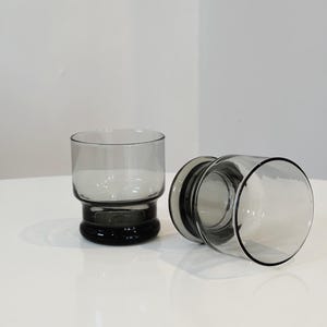 May include: Two gray-tinted drinking glasses. One glass is upright, the other is lying on its side. The glasses have a cylindrical shape with a thick, dark gray base. The glasses are on a white surface.