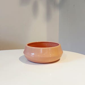 May include: A low-profile, circular ceramic bowl with a smooth, glossy finish in a soft peach color. The bowl features a distinct angled shoulder design that tapers toward the base, creating a wide, open top for versatile use.