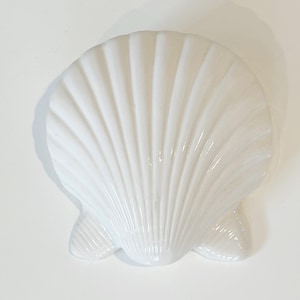 May include: A white, ceramic seashell-shaped dish. The dish has a glossy finish and a detailed, ribbed texture. The design includes three small, rounded feet. This decorative piece is suitable for coastal or nautical-themed decor.