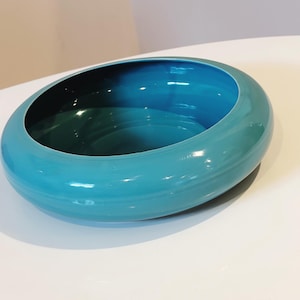 May include: A round, shallow bowl with a glossy, vibrant turquoise finish. The bowl features a wide, smooth, rounded rim that curves inward toward a deep, dark teal interior basin, creating a distinct contrast between the outer and inner surfaces.
