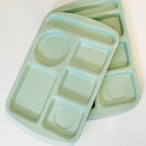 May include: Two light green divided trays with multiple compartments of various shapes, including circles and rectangles. The trays are made of a smooth, solid material and are designed for portion control or serving multiple food items.
