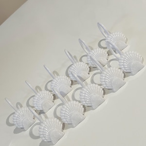 May include: A set of twelve white shower curtain hooks, each featuring a detailed seashell design. The hooks are arranged in two rows, with each piece consisting of a curved metal loop attached to a molded plastic shell ornament in a glossy finish.