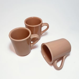 May include: Three brown ceramic coffee mugs with handles. The mugs are arranged on a white surface.