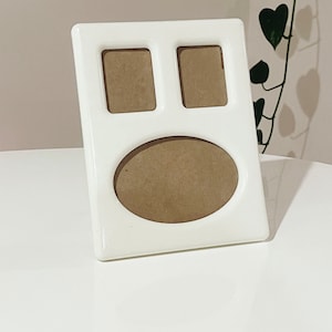 May include: A white picture frame with three openings: two square and one oval. The frame is glossy and sits on a white surface. The openings have a brown backing, ready for photos. A decorative item for displaying memories.