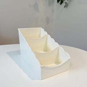 May include: A white, tiered desk organizer with three compartments. The organizer has a scalloped edge design and is made of a smooth, glossy material. The compartments are a light cream color.