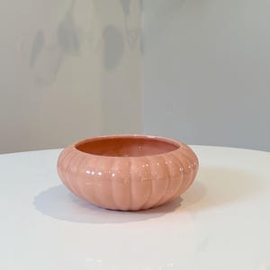 May include: A peach-colored ceramic bowl with a ribbed exterior. The bowl has a rounded shape and sits on a white surface. The interior of the bowl is a slightly darker shade of peach.
