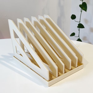 May include: A white plastic magazine holder with five angled compartments. The holder is designed to keep magazines or other documents organized and upright.