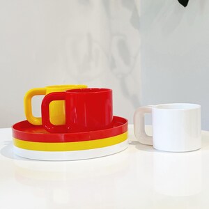 May include: A set of colorful mugs and saucers. One mug is white, while the others are red and yellow. The saucers are red, yellow, and white. The mugs have square handles. The set is on a white surface.