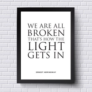 Puede incluir: Impresión tipográfica en blanco y negro con la cita "We are all broken, that's how the light gets in" de Ernest Hemingway. La cita está enmarcada en un marco negro contra una pared de ladrillo blanco.