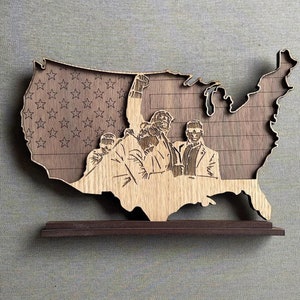 May include: A wooden cutout of the United States of America with a group of people depicted in the center. The left side of the map is a woodgrain pattern with stars, while the right side is a woodgrain pattern with stripes. The map is mounted on a dark brown wooden base.