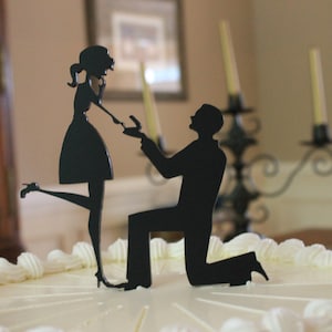 May include: A white wedding cake with a black silhouette cake topper of a couple getting engaged. The man is kneeling and the woman is smiling.