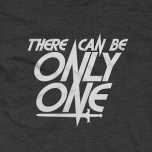 May include: A dark heather grey t-shirt with a white graphic print that reads "There can be only one" in a bold, stylized font. The graphic also includes a sword.