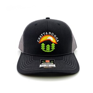 May include: Black and gray trucker hat with a white embroidered logo that says "CHATTANOOGA" with a yellow sun and green trees. The hat has a black and gray Richardson 112 Original Trucker label.