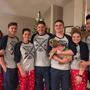 May include: A family of five wearing matching gray and navy blue shirts with the text "Moore 2018 Ski Trip" and a ski graphic. The family is standing in front of a Christmas tree and holding a small brown dog.