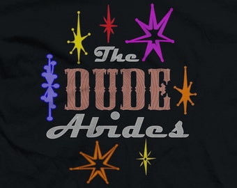 May include: A black t-shirt with a retro design featuring the text "The Dude Abides" in a woodgrain font. The design also includes colorful starburst shapes.