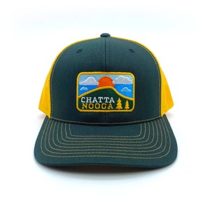 May include: A dark green and yellow trucker hat with an embroidered patch that says "CHATTA NOOGA" and features a mountain scene with a setting sun, clouds, and trees.