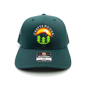 May include: A dark green trucker hat with a white embroidered logo that says "Chattanooga" with a sunset and trees inside the word. The hat has a black mesh back and a white label that says "Richardson 112 Original Trucker One Size".