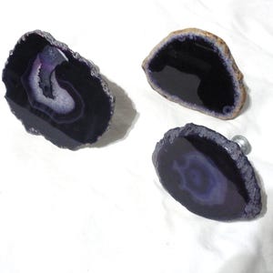 May include: Three decorative cabinet knobs made from polished agate stone. The knobs feature a dark purple color with white and light purple banding. One knob has a silver metal screw attached.