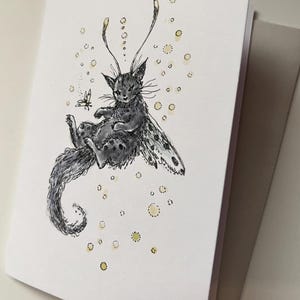 May include: A greeting card featuring a whimsical illustration of a cat with butterfly wings. The cat is gray with black spots and a long, curled tail. A small butterfly hovers nearby, and yellow dots surround the cat. The card is white with a gray envelope.