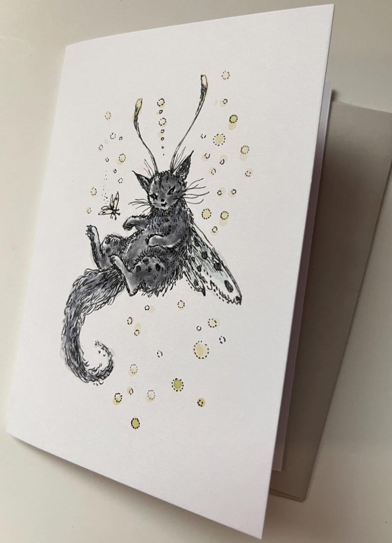 May include: A greeting card featuring a whimsical illustration of a cat with butterfly wings. The cat is gray with black spots and a long, curled tail. A small butterfly hovers nearby, and yellow dots surround the cat. The card is white with a gray envelope.